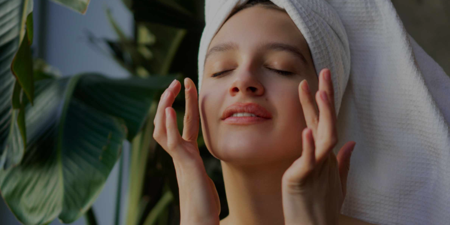 Woman with a towel on her head touching her face with plants in the background